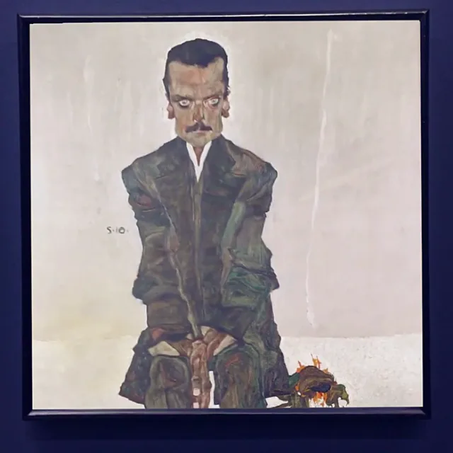 A portrait by Egon Schiele on a dark blue museum wall: a seated man in a dark green suit, hands folded, against a pale ground