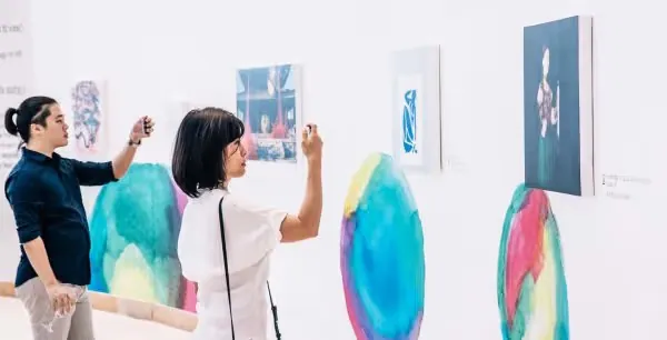 Two visitors photographing colorful artworks on the gallery wall with their phones