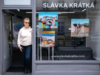 Slávka Krátká standing at the door of her gallery, colorful town paintings displayed in the window