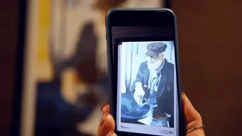 Phone held up to an artwork by Maria Chalela - Puccini, the screen showing the animated painting of a man in a cap