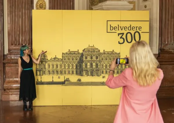 Visitor posing at the yellow Belvedere 300 selfie wall while another photographs her with a phone