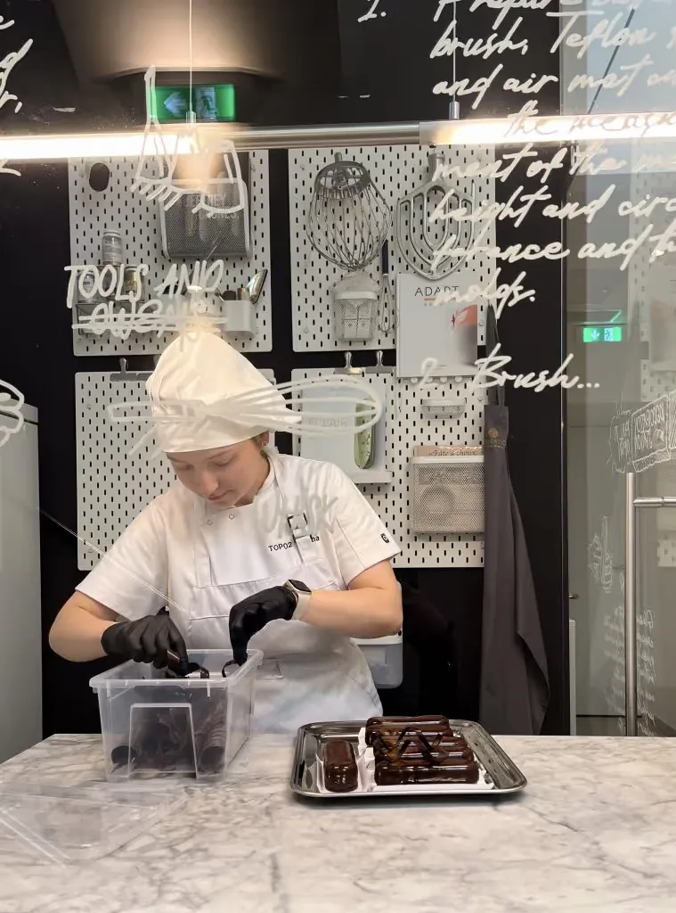 Pastry chef in gloves glazing chocolate eclairs at TOP 02, handwritten mural text on the glass in front