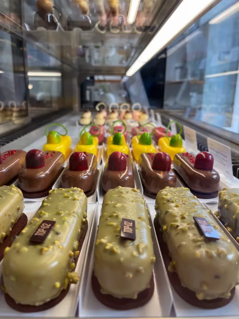 Rows of TOP 02 eclairs in a display case, pistachio, chocolate with cherry and yellow ones with green tops
