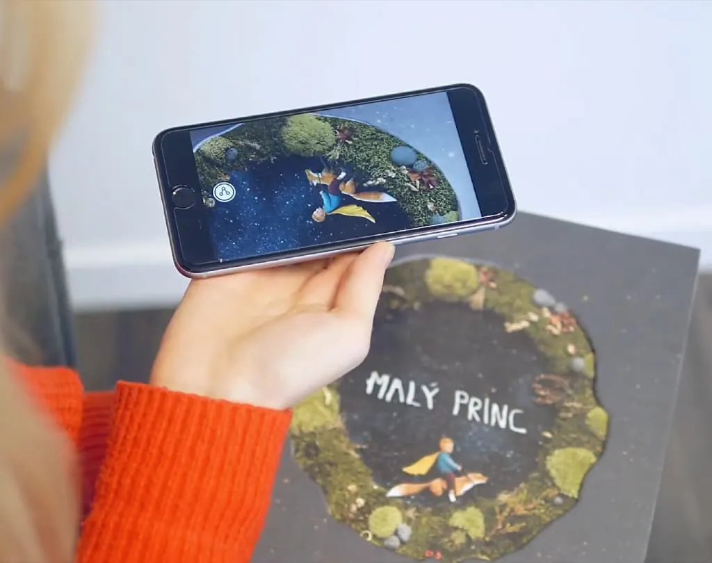 A hand holds a phone over the cover of the Czech edition of The Little Prince, the illustration playing on screen