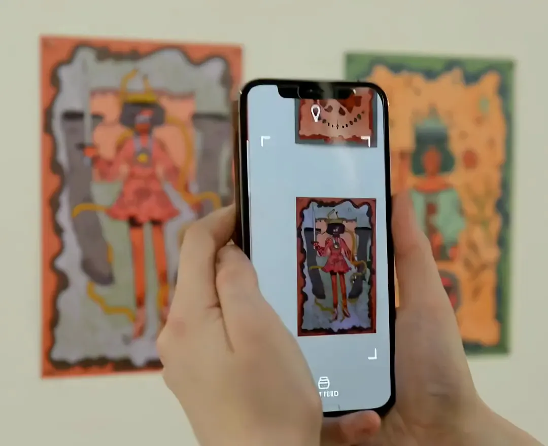 A student holds a phone over a hand-painted tarot card in a gallery; on the screen the card plays its animation
