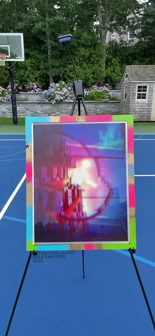 A glitch artwork by Daniel Kramer on an easel outdoors, its AR layer playing on a phone