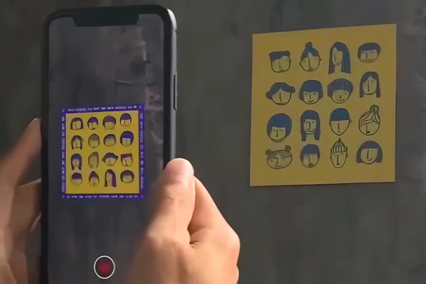 A hand holds a phone up to a print on a concrete wall; on the screen the drawn faces appear inside a blue AR frame