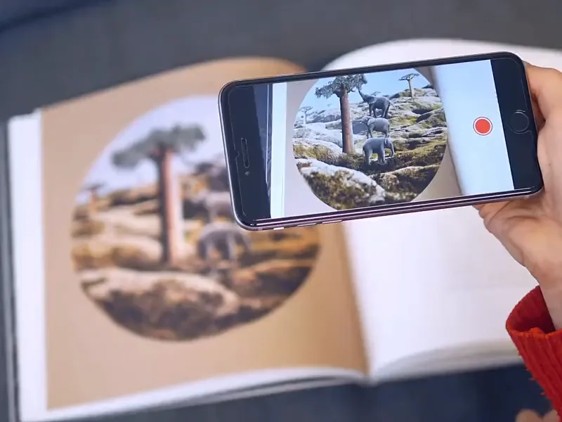 A phone held over an open copy of The Little Prince: the round illustration of baobabs comes to life with elephants walking on the rocks