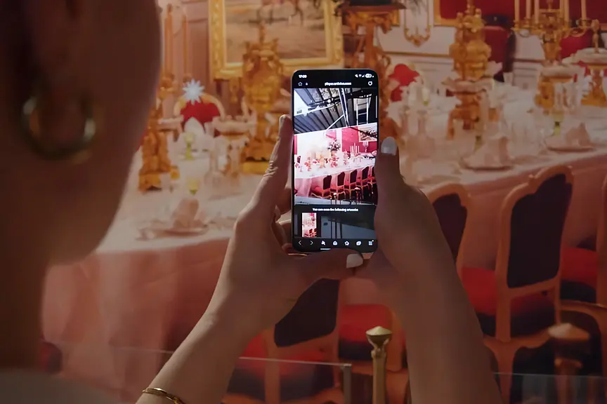 A visitor holds a phone over the imperial dining table at the Sisi Museum; the screen shows the banquet reconstructed with its guests
