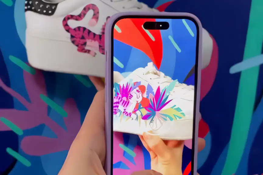 A phone held in front of a white sneaker painted with a pink tiger; on the screen leaves in magenta and green bloom over the shoe
