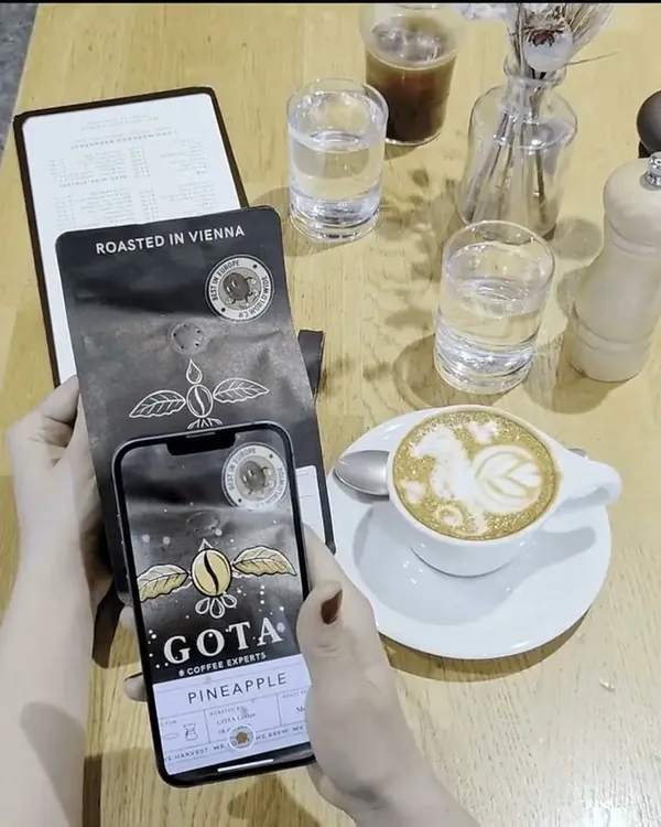 A phone held over a Gota Coffee bag on a café table: on the screen the packaging comes to life