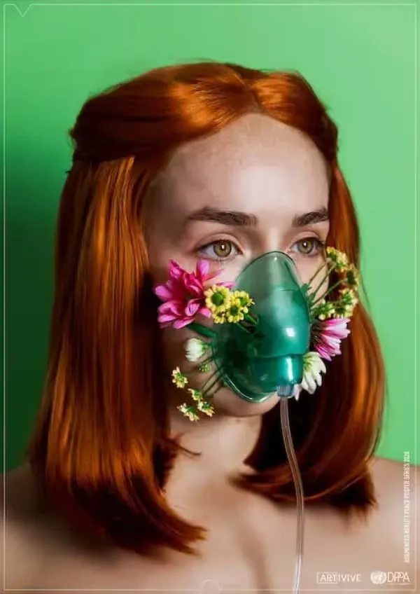 Young woman with red hair wearing a green oxygen mask decorated with pink and yellow flowers against a green background.