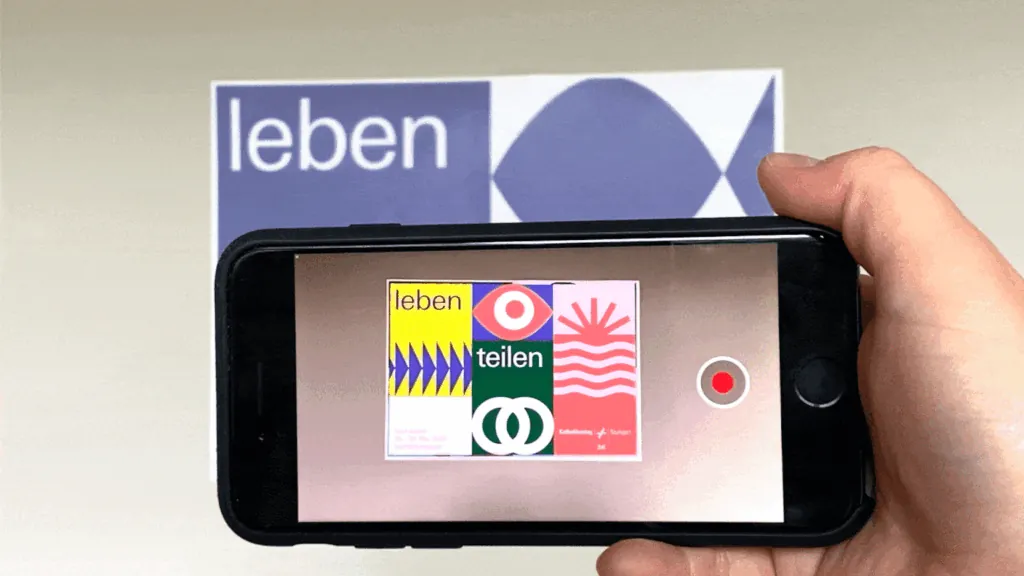 Hand holding a phone with the Artivive app over a leben teilen poster, the screen showing its animated AR layer