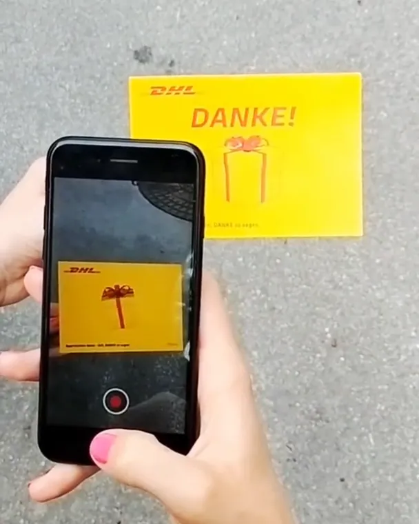A phone scans a yellow DHL thank-you postcard lying on the pavement