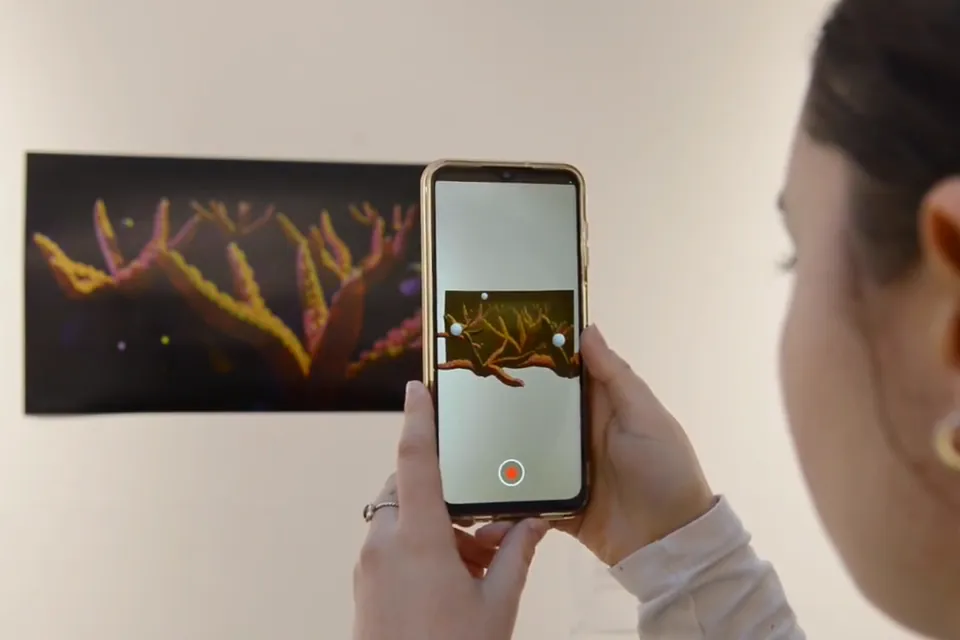 A student holds a phone up to a print of orange tentacles; on screen they grow out of the paper